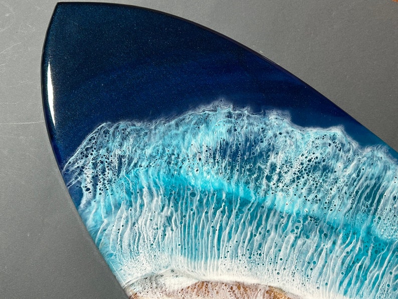 Op de afbeelding: Close-up van een kunstwerk van een surfboard van hars met een blauw en wit golfontwerp. De golf is gemaakt om eruit te zien als brekende oceaan golven met wit schuim.