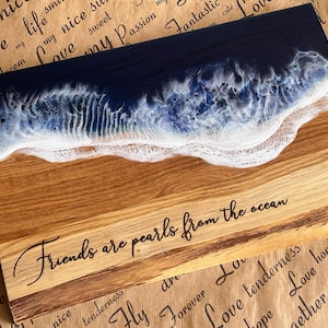 May include: Wooden serving board with a resin ocean design. The top section features a dark blue resin representing the ocean, transitioning to white foam. The bottom section is natural wood with the text "Friends are pearls from the ocean".