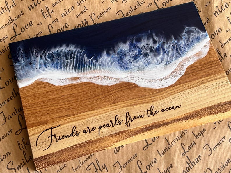 May include: Wooden serving board with a resin ocean design. The top section features a dark blue resin representing the ocean, transitioning to white foam. The bottom section is natural wood with the text "Friends are pearls from the ocean".