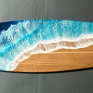 Op de afbeelding: Een houten surfplank serveerschaal met een blauw en wit hars oceaan golf ontwerp. De schaal is gevormd als een surfplank en heeft een natuurlijke houten afwerking.