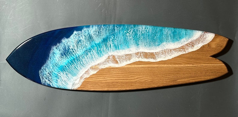 Op de afbeelding: Een houten surfplank serveerschaal met een blauw en wit hars oceaan golf ontwerp. De schaal is gevormd als een surfplank en heeft een natuurlijke houten afwerking.