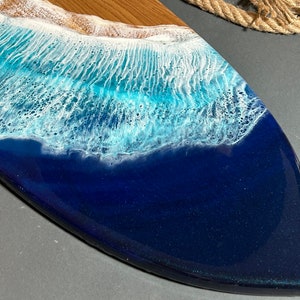 Op de afbeelding: Een houten serveerplank met een blauw en wit hars ocean wave design. De plank is gevormd als een surfboard en heeft een gladde, glanzende afwerking.