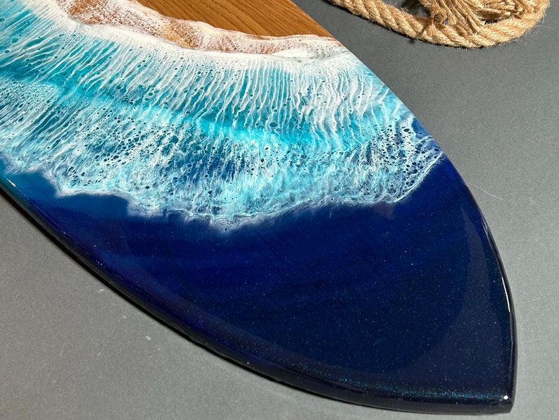 Op de afbeelding: Een houten serveerplank met een blauw en wit hars ocean wave design. De plank is gevormd als een surfboard en heeft een gladde, glanzende afwerking.
