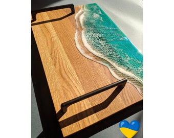 Op de afbeelding: Een rechthoekig houten dienblad met een natuurlijke houtnerfafwerking en twee zwarte metalen handgrepen. Eén kant van het blad is versierd met een glanzende harsinleg die het uiterlijk van turquoise oceaangolven met wit schuim nabootst.
