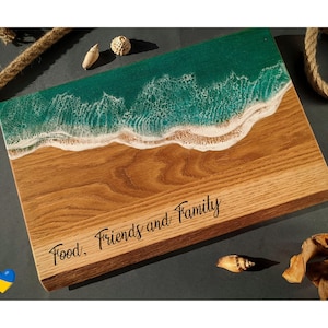 May include: Wooden serving board with a beach-themed design. The top features a turquoise resin ocean with white foamy waves, transitioning to a sandy-colored wood base. The words "Food, Friends and Family" are inscribed on the wood.