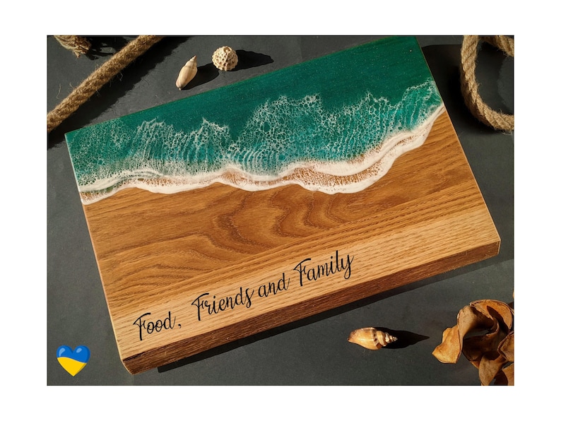 May include: Wooden serving board with a beach-themed design. The top features a turquoise resin ocean with white foamy waves, transitioning to a sandy-colored wood base. The words "Food, Friends and Family" are inscribed on the wood.