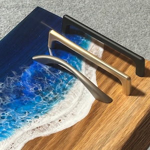 May include: Three cabinet pulls in silver, gold, and black are displayed on a wooden surface with a resin art design of a beach scene. The pulls are straight with a curved handle.