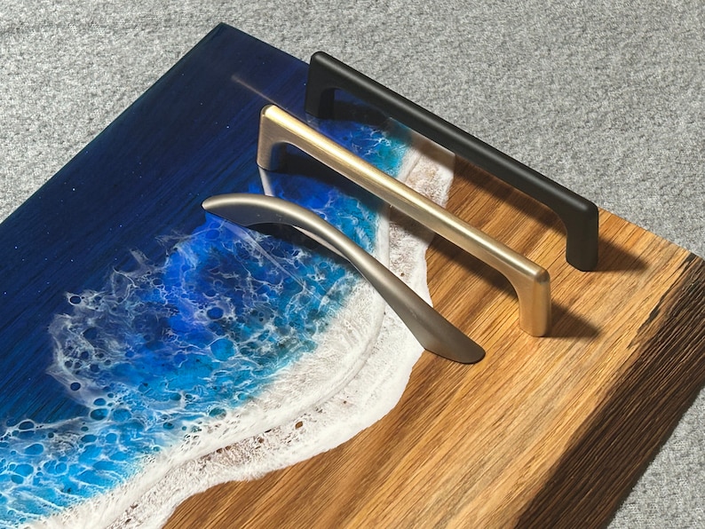 May include: Three cabinet pulls in silver, gold, and black are displayed on a wooden surface with a resin art design of a beach scene. The pulls are straight with a curved handle.