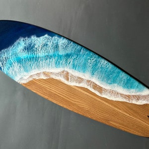 Op de afbeelding: Een houten surfboard met een blauw en wit harsgolvenontwerp. De golf is gemaakt om eruit te zien als een brekende oceaan golf met wit schuim.
