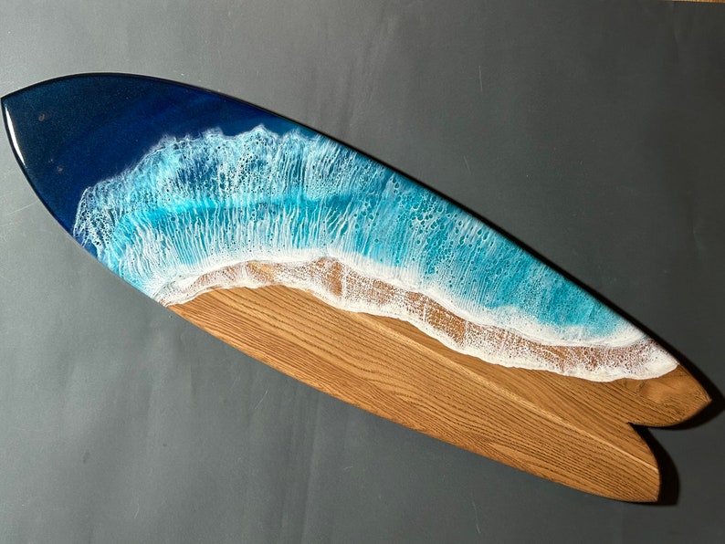Op de afbeelding: Een houten surfboard met een blauw en wit harsgolvenontwerp. De golf is gemaakt om eruit te zien als een brekende oceaan golf met wit schuim.