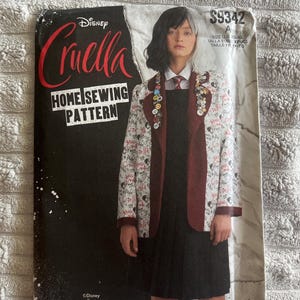 May include: A Disney Cruella home sewing pattern envelope by Simplicity, model S9342. The cover features a person wearing a white blazer with a black text print, a dark red lapel, and a black dress. The pattern is for sizes XS to XL.