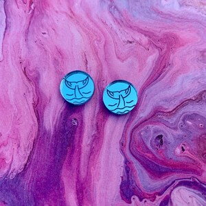 K&ouml;nnte beinhalten: Zwei runde, hellblaue Ohrstecker. Jeder Ohrring zeigt eine schwarze Strichzeichnung einer Walflosse &uuml;ber wellenf&ouml;rmigen Linien. Die Ohrringe sind vor einem rosa-violett marmorierten Hintergrund platziert.