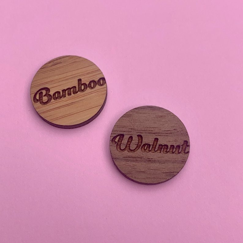 K&ouml;nnte beinhalten: Zwei runde Holzst&uuml;cke, eines hellbraun mit der Gravur "Bamboo", das andere dunkelbraun mit der Gravur "Walnut". Die St&uuml;cke liegen auf einem rosa Hintergrund. Das Bambusst&uuml;ck hat eine sichtbare Holzmaserung.