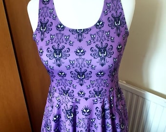 Puede incluir: Un vestido morado sin mangas con un patrón repetido de fantasmas negros y una silueta de mansión. El vestido tiene una falda acampanada y un escote redondo.