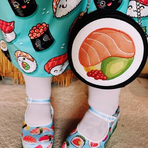 May include: A pair of white platform shoes with a blue and green background and a variety of sushi designs. The shoes are worn by a person wearing a blue and white dress with a sushi print. The person is also carrying a black round purse with a sushi design.