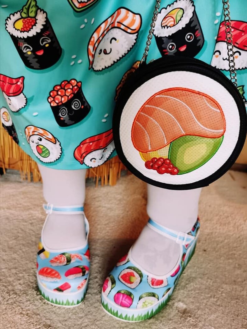May include: A pair of white platform shoes with a blue and green background and a variety of sushi designs. The shoes are worn by a person wearing a blue and white dress with a sushi print. The person is also carrying a black round purse with a sushi design.