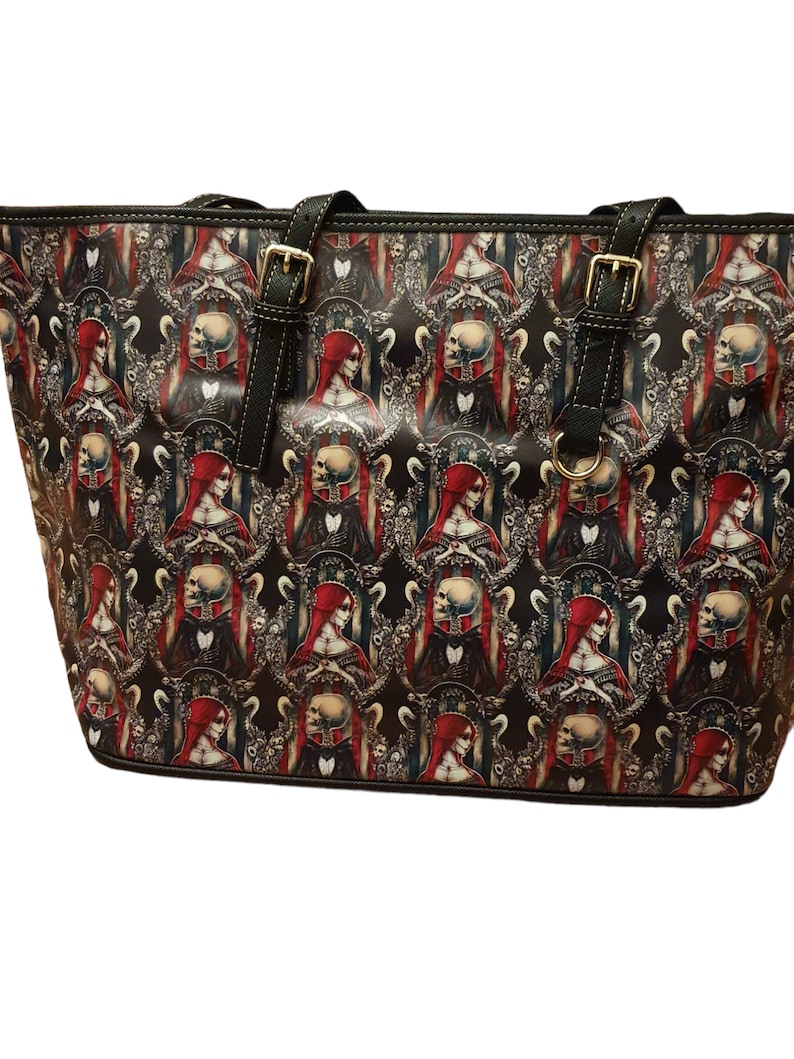 Puede incluir: Una bolsa de mano negra con un patr&oacute;n repetido de una mujer con cabello rojo y una calavera. El patr&oacute;n est&aacute; sobre un fondo negro con detalles dorados.