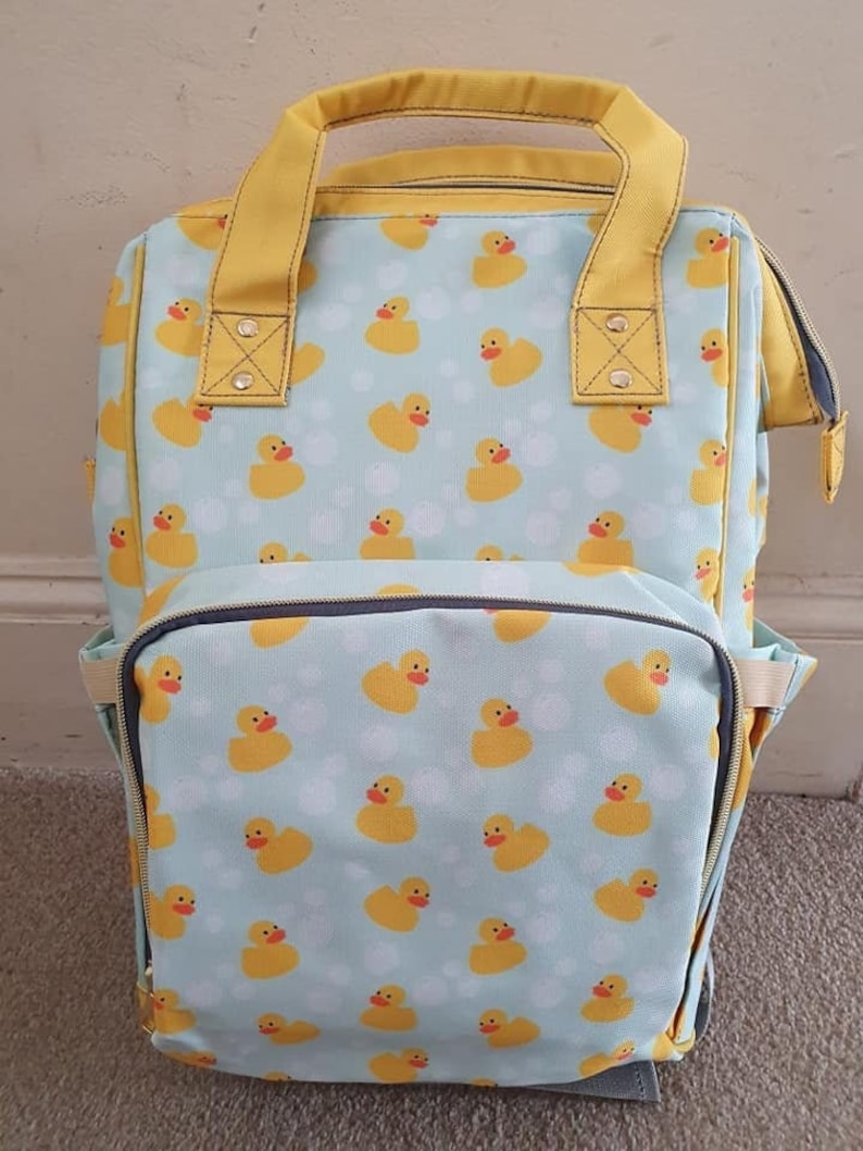 May include: A light blue and yellow backpack with a pattern of rubber duckies. The backpack has a large main compartment and a smaller front pocket. The straps are yellow.