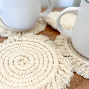 Puede incluir: Primer plano de dos tazas de cerámica blanca con bordes gris oscuro, sobre posavasos de macramé color crema. Los posavasos tienen un diseño en espiral y bordes con flecos. Las tazas están sobre una superficie de madera. Los posavasos miden unos 10 cm de diámetro.