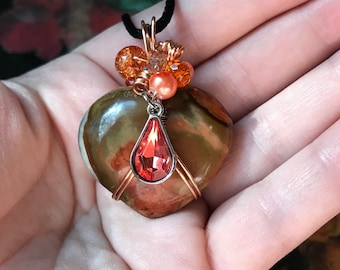 May include: A heart-shaped pendant made of brown and green agate with copper wire wrapping. The pendant is adorned with a teardrop-shaped red crystal and a cluster of orange and clear beads.