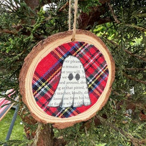 K&ouml;nnte beinhalten: Runder Weihnachtsbaumschmuck aus Holz mit rotem und schwarzem Karomuster. Der Schmuck hat eine ausgeschnittene Silhouette, mit Text darin. Er h&auml;ngt an einer Schnur aus Naturfaser.