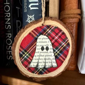 K&ouml;nnte beinhalten: Ein runder Holzschmuck mit einem rot-schwarz-gelben Karomuster. In der Mitte befindet sich ein Ausschnitt mit Text aus einem Buch. Der Schmuck wird an einer nat&uuml;rlichen Schnur aufgeh&auml;ngt, ideal f&uuml;r Feiertagsdekorationen.