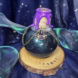 May include: A dark blue, round potion bottle with a purple, dripped-wax top and a gold chain with a witch hat charm. The bottle sits on a wooden slice with a crescent moon design. The background is a dark blue fabric with stars.