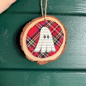 K&ouml;nnte beinhalten: Runder Weihnachtsbaumschmuck aus Holz mit einem Einsatz aus rot-schwarz-wei&szlig;em Karostoff. Eine wei&szlig;e Geisterform ist aus dem Stoff ausgeschnitten. Der Schmuck wird an einer Schnur aufgeh&auml;ngt.