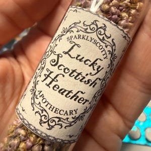 May include: A clear glass vial filled with small, dried purple and beige heather flowers. A white paper label is wrapped around the center, featuring black decorative scrollwork and the text: Sparklyscotty Lucky Scottish Heather Apothecary.