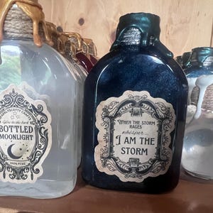 May include: A clear glass bottle with gold wax drips and a label reading BOTTLED MOONLIGHT, next to a dark blue bottle with green wax drips and a label reading WHEN THE STORM RAGES whisper... I AM THE STORM, and a small clear bottle with a ghost charm.