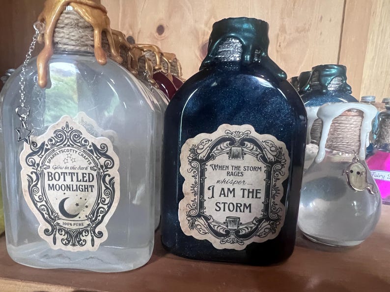 May include: A clear glass bottle with gold wax drips and a label reading BOTTLED MOONLIGHT, next to a dark blue bottle with green wax drips and a label reading WHEN THE STORM RAGES whisper... I AM THE STORM, and a small clear bottle with a ghost charm.