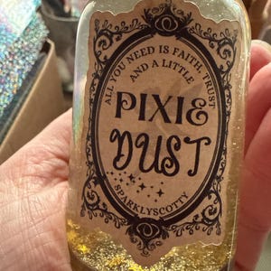 Könnte beinhalten: Eine kleine Glasflasche gefüllt mit goldenem Glitzer, beschriftet mit "Pixie Dust". Das Etikett zeigt verzierte schwarze Schnörkel und den Text "All you need is faith and a little trust". Die Flasche wird in einer Hand gehalten.