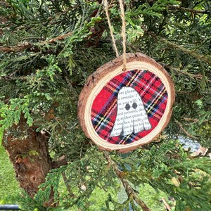 K&ouml;nnte beinhalten: Ein runder Weihnachtsbaumschmuck aus Holz mit einem Einsatz aus rot-schwarz kariertem Stoff. Der Anh&auml;nger hat einen nat&uuml;rlichen Holzrand und wird an einer Schnur aufgeh&auml;ngt. Text ist auf dem Stoffeinsatz sichtbar.