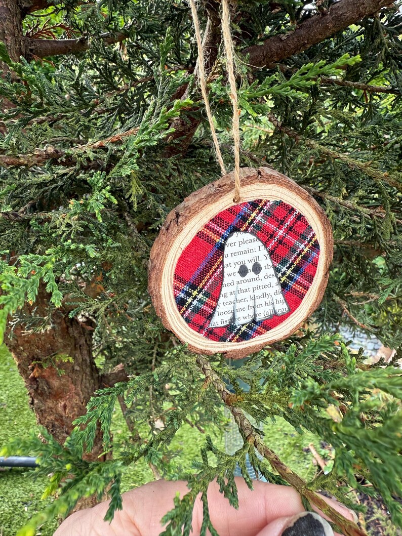 K&ouml;nnte beinhalten: Ein runder Weihnachtsbaumschmuck aus Holz mit einem Einsatz aus rot-schwarz kariertem Stoff. Der Anh&auml;nger hat einen nat&uuml;rlichen Holzrand und wird an einer Schnur aufgeh&auml;ngt. Text ist auf dem Stoffeinsatz sichtbar.