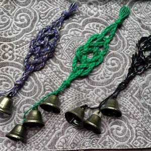 May include: Three decorative bell ornaments in blue, green, and black. Each ornament features a braided design and multiple small brass bells. The bells are suspended from the braided section by a thin cord.