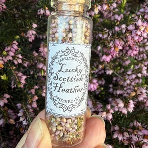 May include: A clear glass vial with a cork stopper filled with dried pink and white heather flowers. A white rectangular label on the front features black decorative borders and the text: Sparklyscotty Lucky Scottish Heather Apothecary.