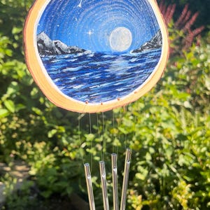 May include: A circular wood slice wind chime featuring a hand-painted night scene of a full moon over a dark blue ocean with rocky mountains on either side. Four silver metal cylindrical chimes hang from the wood slice by thin clear strings.