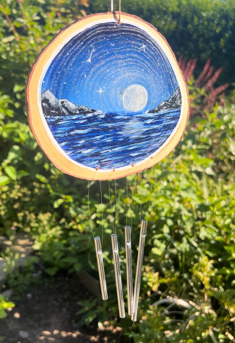 May include: A circular wood slice wind chime featuring a hand-painted night scene of a full moon over a dark blue ocean with rocky mountains on either side. Four silver metal cylindrical chimes hang from the wood slice by thin clear strings.