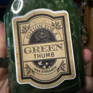 May include: A green, translucent bottle filled with a shimmering liquid. A rectangular label features a floral illustration and the text: POTION FOR A GREEN THUMB MAX STREGNTH. The bottle is sealed with a wax-like substance at the top opening.