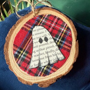 K&ouml;nnte beinhalten: Ein Holzscheiben-Ornament mit rotem und schwarzem Karostoff. Ein wei&szlig;er Geist mit schwarzen Augen ist auf dem Stoff zentriert. Das Ornament hat eine Aufh&auml;ngung aus Juteschnur.