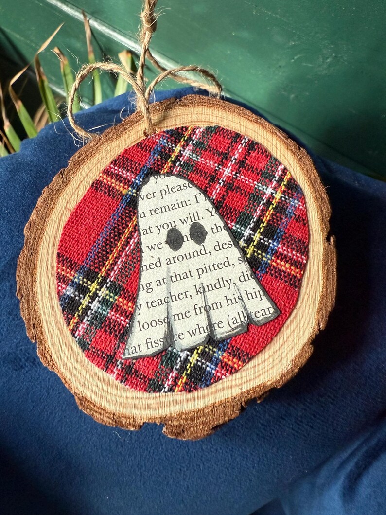 K&ouml;nnte beinhalten: Ein Holzscheiben-Ornament mit rotem und schwarzem Karostoff. Ein wei&szlig;er Geist mit schwarzen Augen ist auf dem Stoff zentriert. Das Ornament hat eine Aufh&auml;ngung aus Juteschnur.