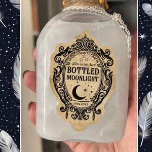 May include: A clear glass bottle filled with a shimmering white liquid, featuring a decorative tan label with black text reading "SPARKLYSCOTTY CRAFTS Glow in the dark BOTTLED MOONLIGHT 100% PURE" and a crescent moon icon. The bottle neck is wrapped in twine.