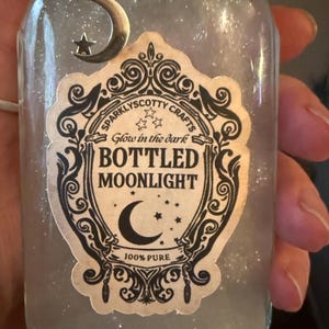 May include: A clear glass bottle filled with a shimmering, translucent liquid. A silver crescent moon and star charm hangs from the neck. The label features ornate black scrollwork and text reading: Sparklyscotty Crafts, Glow in the dark, BOTTLED MOONLIGHT, 100% PURE.
