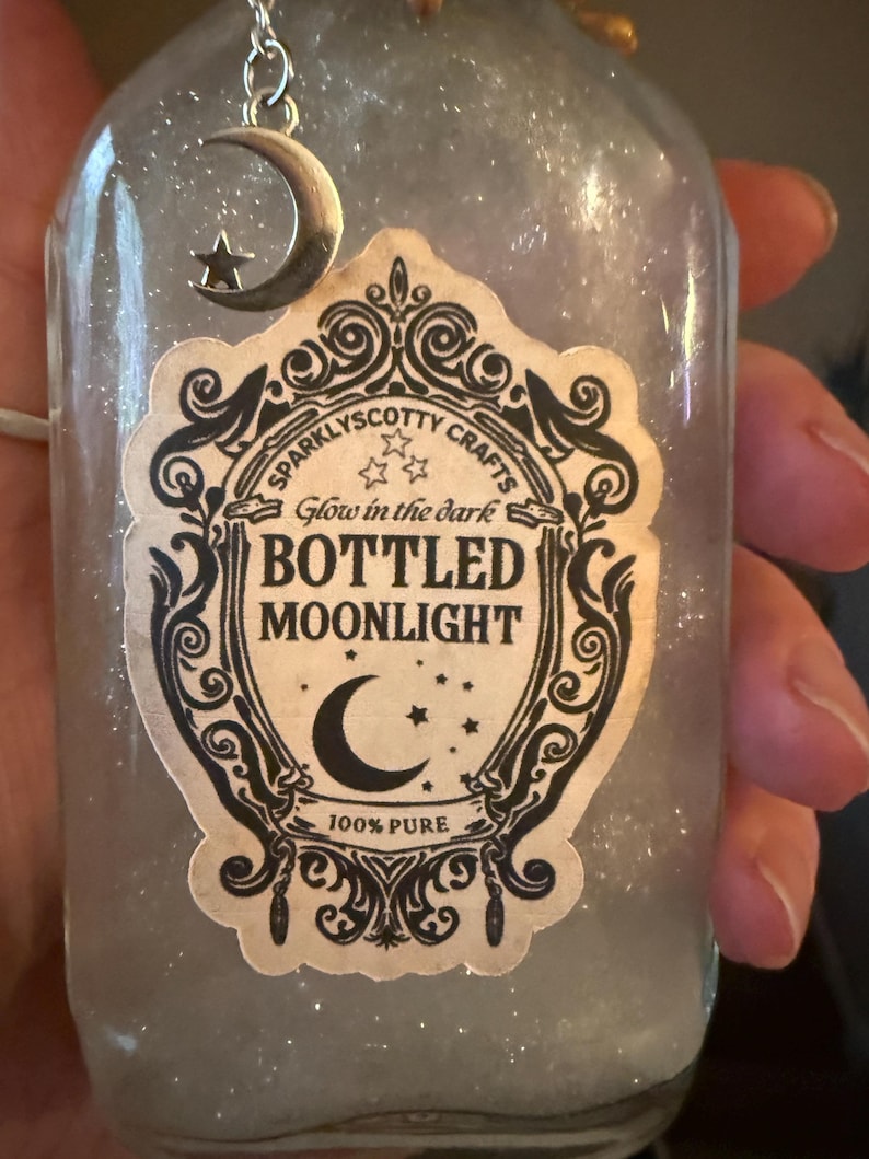 May include: A clear glass bottle filled with a shimmering, translucent liquid. A silver crescent moon and star charm hangs from the neck. The label features ornate black scrollwork and text reading: Sparklyscotty Crafts, Glow in the dark, BOTTLED MOONLIGHT, 100% PURE.