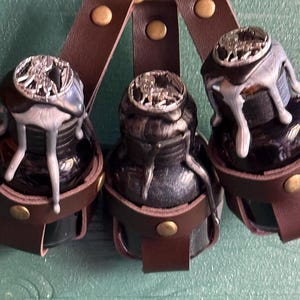 Może przedstawiać: Trzy czarne kałamarze ze srebrnymi wieczkami i brązowymi skórzanymi paskami. Butelki mają metaliczny, ociekający efekt. Paski są przymocowane do ozdobnego zapięcia w kolorze mosiądzu. Unikalne artykuły papiernicze.