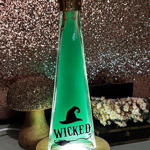 Puede incluir: Una botella de vidrio verde con un sombrero de bruja y la palabra "WICKED" en ella. La botella está iluminada desde abajo, creando un brillo cálido. La botella está sentada sobre una base de madera.