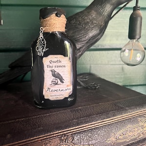 May include: A black glass bottle with a label reading "Quoth the raven Nevermore" and a raven illustration. The bottle has a twine-wrapped top and a charm. A black raven statue and a light bulb are in the background.