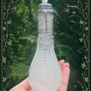 May include: A pear-shaped, frosted glass bottle with a silver-colored top and decorative chain accents. The bottle features a clear, faceted stopper and a dangling blue bead. The design evokes a mystical or fantasy theme.