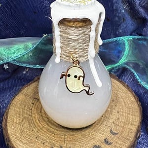 Può includere: Una bottiglia di vetro decorativa con un liquido bianco lattiginoso all'interno. La bottiglia è sigillata con un tappo di sughero, spago e cera bianca. Un ciondolo a forma di fantasma dorato pende dallo spago. La bottiglia è su una fetta di legno.
