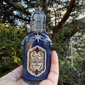Peut inclure: Une bouteille en verre bleu foncé, recouverte de paillettes, avec un dessus en cire argentée dégoulinante. La bouteille porte une étiquette indiquant "Bottled Starlight" et est ornée d'une étoile et d'une chaîne. La bouteille est tenue dans une main.